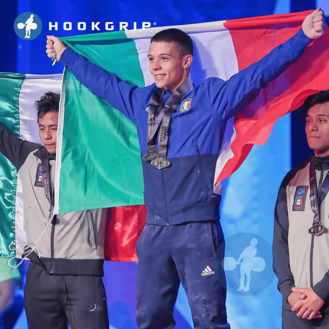 Sergio Massidda, atleta italiano di sollevamento pesi, alza orgogliosamente la bandiera italiana sul podio dopo una vittoria in gara. DRACONESChronicles